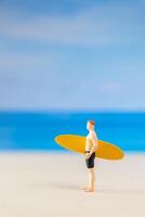 miniatura pessoas homem dentro uma roupa de banho, e segurando uma amarelo prancha de surfe em a de praia foto