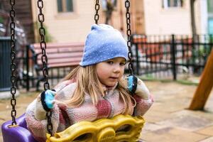 uma jovem menina senta em uma balanço dentro uma Parque infantil, dela expressão é pensativo e introspectivo. ela desgasta uma aconchegante, casual roupa, caloroso chapéu e luvas. foto