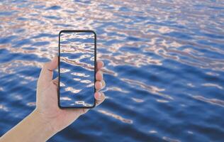jovem homem mão levando foto do lindo luz reflexão em mar superfície com Smartphone dentro pôr do sol Tempo