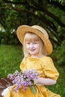 uma pequeno menina dentro uma amarelo vestir e Palha chapéu vestindo uma ramalhete do lilases. uma andar dentro uma Primavera parque, florescendo lilases. foto