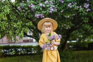 uma pequeno menina dentro uma amarelo vestir e Palha chapéu vestindo uma ramalhete do lilases. uma andar dentro uma Primavera parque, florescendo lilases. foto