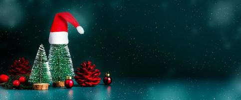 Feliz Natal e Feliz Ano Novo a neve caindo com a árvore de Natal e o chapéu vermelho de Papai Noel em fundo verde escuro. Espaço de maquete de baner para exibição de produto ou projeto ou texto foto