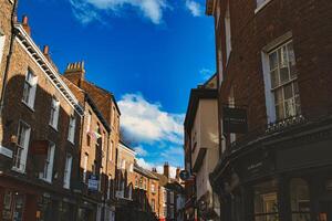 encantador europeu rua cena com histórico tijolo edifícios debaixo uma Claro azul céu com fofo nuvens, exibindo arquitetônico detalhes e local negócios dentro Iorque, norte yorkshire, Inglaterra. foto