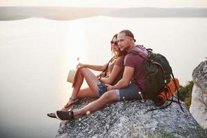 casal abraçando com mochila sentado no topo da montanha rochosa, apreciando a vista da costa de um rio ou lago. viajando ao longo das montanhas e da costa, conceito de liberdade e estilo de vida ativo foto