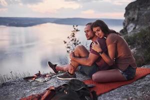 casal abraçando com mochila sentado perto do fogo no topo da montanha, apreciando a vista da costa de um rio ou lago. viajando ao longo das montanhas e da costa, conceito de liberdade e estilo de vida ativo foto