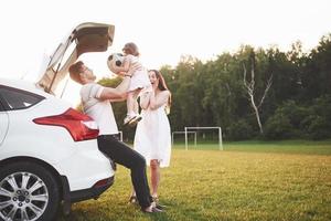 muito jovem casal e sua filha estão descansando na natureza. a mãe, o pai e a filha estão sentados no porta-malas do carro aberto foto