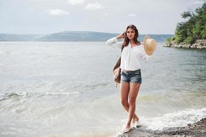 uma linda jovem com um chapéu e uma mochila caminha divertidamente à beira da água. um dia quente de verão é uma ótima época para aventura e aventura na natureza foto
