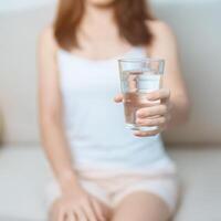 feliz mulher segurando água vidro, fêmea bebendo puro água em sofá às lar. saudável, refresco, estilo de vida e diariamente rotina conceito foto