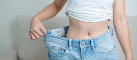 jovem fino fêmea vestindo grande ou grande demais jeans, mulher mostrar saudável forma depois de peso perda. fazer dieta, exercício, corpo em forma e lipoaspiração cirurgia conceito foto