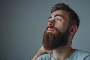 ai gerado bonito jovem barbudo homem é guardando mão em barba e olhando longe em uma cinzento fundo foto