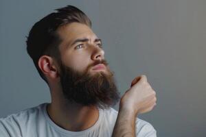 ai gerado bonito jovem barbudo homem é guardando mão em barba e olhando longe em uma cinzento fundo foto