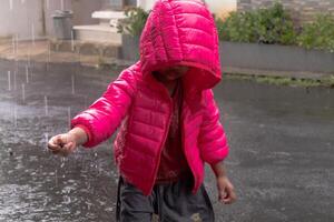 a ásia pequeno menina vestindo uma Rosa Jaqueta e jogando dentro a chuva dentro frente do a casa foto