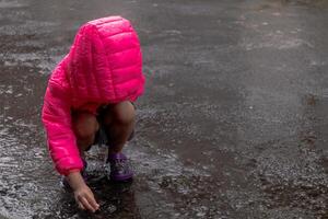 a ásia pequeno menina vestindo uma Rosa Jaqueta e jogando dentro a chuva em a asfalto estrada foto