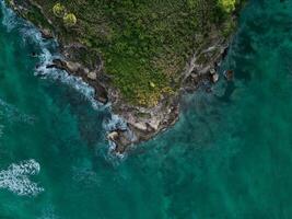 aéreo. Sombrio turquesa água com branco espumoso ondas e verde rochoso costa foto