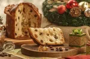 uma fatia de panetone e cubos de frutas cristalizadas em uma tábua de madeira com enfeites de natal foto