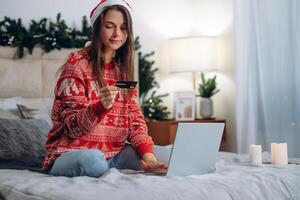 Natal conectados compras, vendas e descontos promoções durante inverno feriados, conectados compras às lar. fêmea em cama com computador portátil com crédito cartão e Natal decorações foto