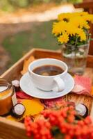 uma copo do chá ou café em uma de madeira bandeja contra a fundo do caído folhas, vela, outono temporada, ainda vida com folhas, flores e castanhas foto