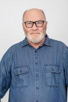 retrato do a atraente grisalho homem com óculos vestindo uma jeans camisa isolado em uma cinzento fundo. barbudo avô do a europeu tipo. foto