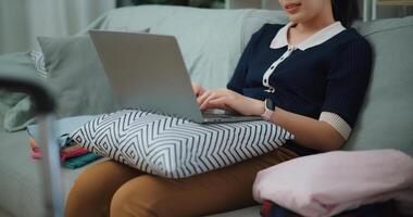 seletivo foco mãos do ásia adolescente mulher sentado em sofá usando computador portátil para preparar reserva hotel e avião bilhete para viagem. mochileiro viagem conceito. foto