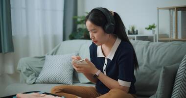 seletivo foco do ásia adolescente mulher vestem sem fio fones de ouvido sentado em sofá fazer lista de controle do coisas para pacote para viagem, preparação viagem mala de viagem às lar. foto