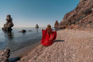 uma mulher dentro uma vermelho vestir carrinhos em uma de praia com uma rochoso litoral dentro a fundo. a cena é sereno e pacífico, com a mulher vermelho vestir contrastante contra a natural elementos do a de praia. foto