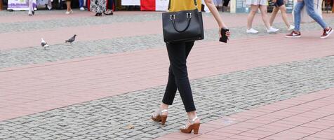 mulher vestindo uma Preto Bolsa e segurando uma inteligente telefone dentro dela mão é caminhando em a vista da rua de a costas foto