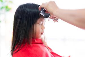 fofa ásia menina sentado imóvel com olhos fechadas e vestindo uma vermelho cabelo corte capa com uma barbeiro, mãos do a barbeiro segurando uma pente e tesouras para cortar a frente cabelo com uma pequeno criança. foto