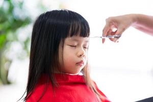 ásia menina é sentado e tendo dela cabelo cortar de barbeiro, criança é vestindo vermelho cabelo corte capa para evita cabelo partículas a partir de degola para dela roupas e corpo, criança senta silenciosamente com dela olhos fechado. foto