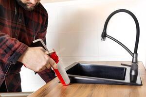 uma homem instala uma Preto pedra Pia para dentro uma cozinha bancada. trabalhador selos acima a cozinha Pia com uma selante usando uma construção selagem arma de fogo foto