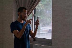 jovem ásia homem em pé em janela enquanto apontando dele Smartphone foto