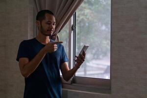 sentindo-me feliz do jovem ásia homem dentro azul camiseta e apontando para inteligente telefone ao lado do janela foto