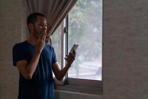 jovem ásia homem dentro azul camiseta em pé em janela fechar dele boca. pegue surpreso e sentir feliz de dele Smartphone foto