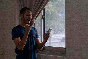 animado jovem ásia homem em pé ao lado do janela com inteligente telefone foto