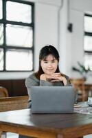 contemplativo mulher descansos dela queixo em dela mãos enquanto olhando às dela computador portátil tela dentro a acolhedor espaço do uma cafeteria. foto