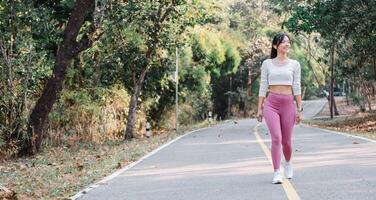 mulher dentro uma branco cortada topo e Rosa perneiras caminhando em uma parque caminho com verde árvores em a lados. foto