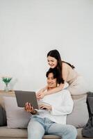 sorridente ásia casal é confortavelmente noivando com uma computador portátil, exibindo uma momento do compartilhado descoberta e prazer em seus sofá. foto