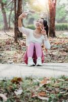 alegre jovem mulher sentado em uma caminho dentro uma iluminado pelo sol parque leva uma selfie, piscando uma Paz placa. foto