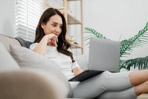 pensativo jovem mulher goza a conforto do dela moderno vivo quarto enquanto navegando em dela computador portátil, incorporando uma relaxado trabalho a partir de casa vibe. foto