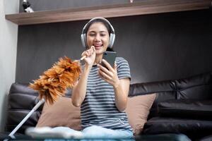 casa limpeza com Diversão. feliz jovem ásia dona de casa cantando música durante limpar, usando pena espanador Como microfone, desfrutando doméstico trabalhar. jovem mulher dançando e limpeza dentro vivo quarto foto