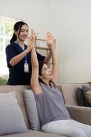 feliz Senior mulher fazendo exercício às casa com ásia fêmea fisioterapeuta. velho aposentado senhora fazendo alongamento braços às casa com a Socorro do uma pessoal fisioterapeuta durante uma reabilitação sessão foto