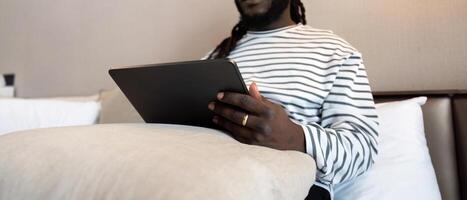 sorridente jovem africano americano homem usando digital tábua controlo remoto trabalho sentado em a cama às lar. conceito conectados tecnologia comunicação foto