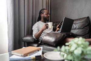 jovem africano americano homem segurando computador portátil relaxante sentado em sofá trabalhando controlo remoto às casa foto