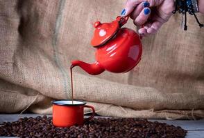 mão com unhas azuis com bule de metal vermelho fazendo café em copo de metal vermelho foto