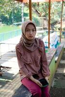 javanese mulher dentro hijab é sentado dentro a parque com uma sorridente expressão, verão período de férias conceito. foto