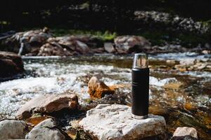 acampamento de a água dentro uma montanha rio, uma conceptual cenário do uma caminhada garrafa térmica para chá, uma garrafa para quente bebidas, não pessoas, uma tormentoso corrente do água. foto