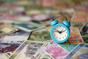 pequeno alarme relógio em muitos notas do diferente moeda. fundo do Tempo e dinheiro foto