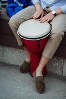 uma branco cara tocam uma tambor, uma rua jazz músico em uma djemba, uma vermelho musical instrumento, Castanho macassins, uma Assistir em dele braço. foto