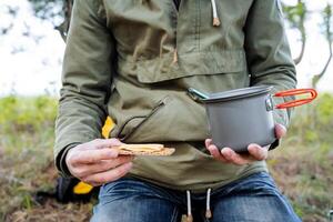 café da manhã do uma turista dentro a floresta, uma pessoa janta em uma sanduíche, uma mão detém uma pão do pão com queijo, uma Panela do mingau, turista utensílios, acampamento Comida dentro natureza, caminhada Comida foto