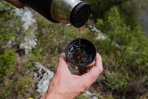 uma homem derrama chá a partir de uma garrafa térmica para dentro uma caneca tiro fechar-se, a conceito do quente bebidas dentro natureza dentro a acampamento, uma copo com bolhas do água a partir de chá. foto