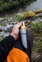 acampamento turista faca bushcraft para sobrevivência dentro a floresta. uma homem segurando uma faca dentro dele mão dentro uma caso liderar em dele joelho. cinzento plástico bainha. foto